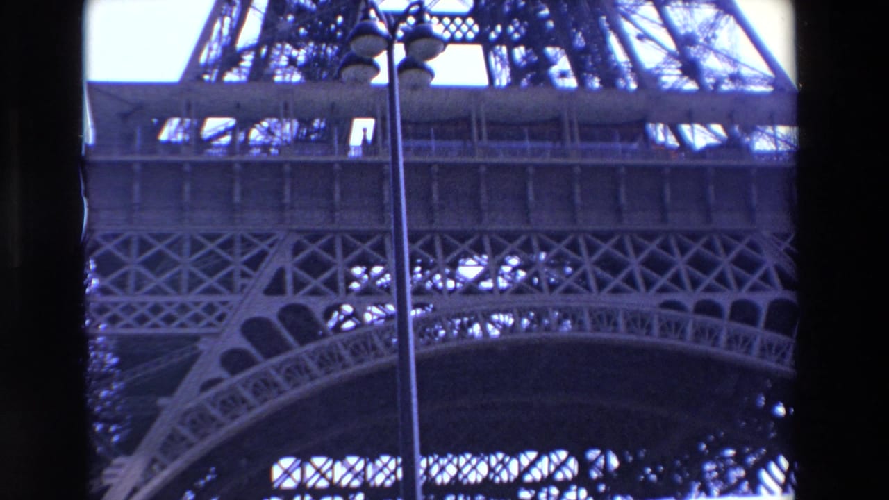 1969 Paris 8mm frame with the Eiffel Tower platform, crisscross iron lattice, lower arch, bright sky, and a lamppost cutting vertically through the view.