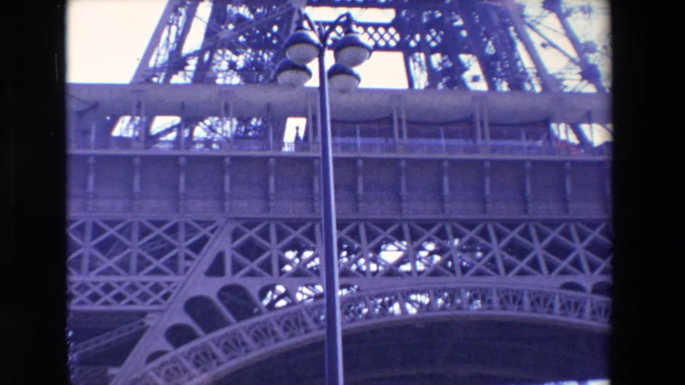 1969 Paris 8mm frame showing a closer upward view of the Eiffel Tower platform and geometric iron lattice, with a foreground lamppost and dark film-edge border.