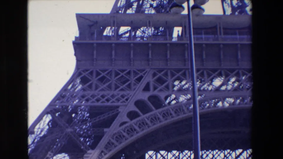 1969 Paris 8mm frame showing the Eiffel Tower lower arch and dense lattice structure from below, with a vertical lamppost and bright sky in the composition.