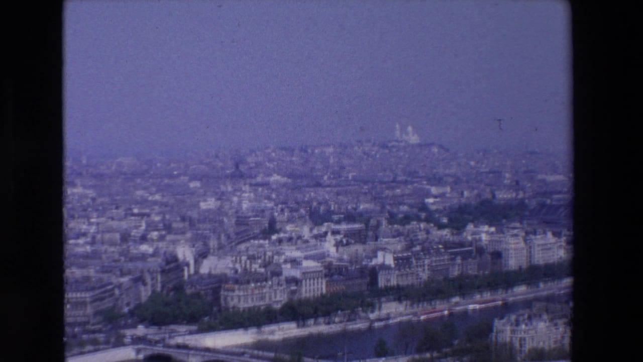 Hazy elevated view over Paris with the Seine, a bridge, dense buildings, and Sacré-Cœur in the distance.