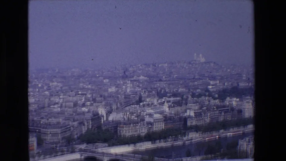 Vintage 8mm frame of Paris in 1969, showing a broad urban view over the riverfront with Sacré-Cœur appearing faintly beyond the city blocks.