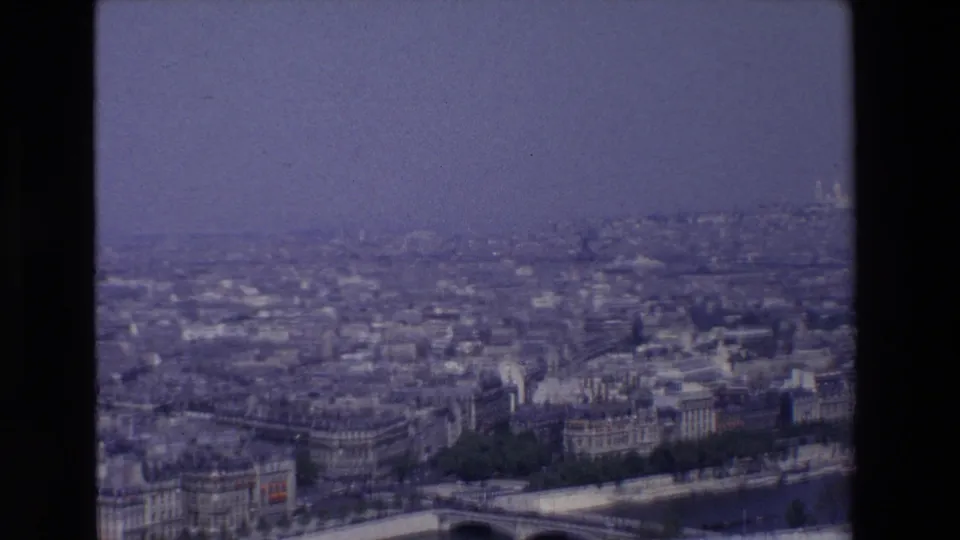 1969 8mm view over Paris, France, showing dense rooftops, riverside buildings, bridge arches, and a hazy skyline framed by the dark edge of the film gate.