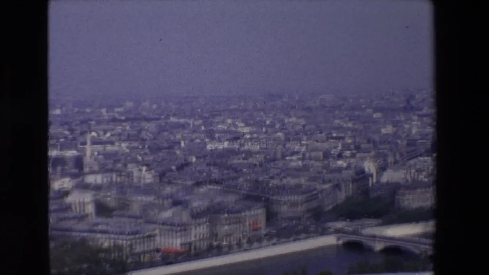 1969 8mm aerial-style view of Paris with soft focus, a river edge, bridge structure, clustered rooftops, and muted winter trees near the lower frame.