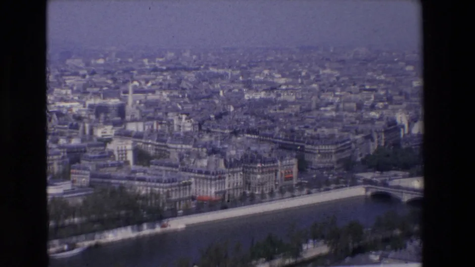 1969 8mm Paris riverfront panorama showing the Seine River, a bridge, dense urban blocks, bare trees along the embankment, and a hazy skyline.