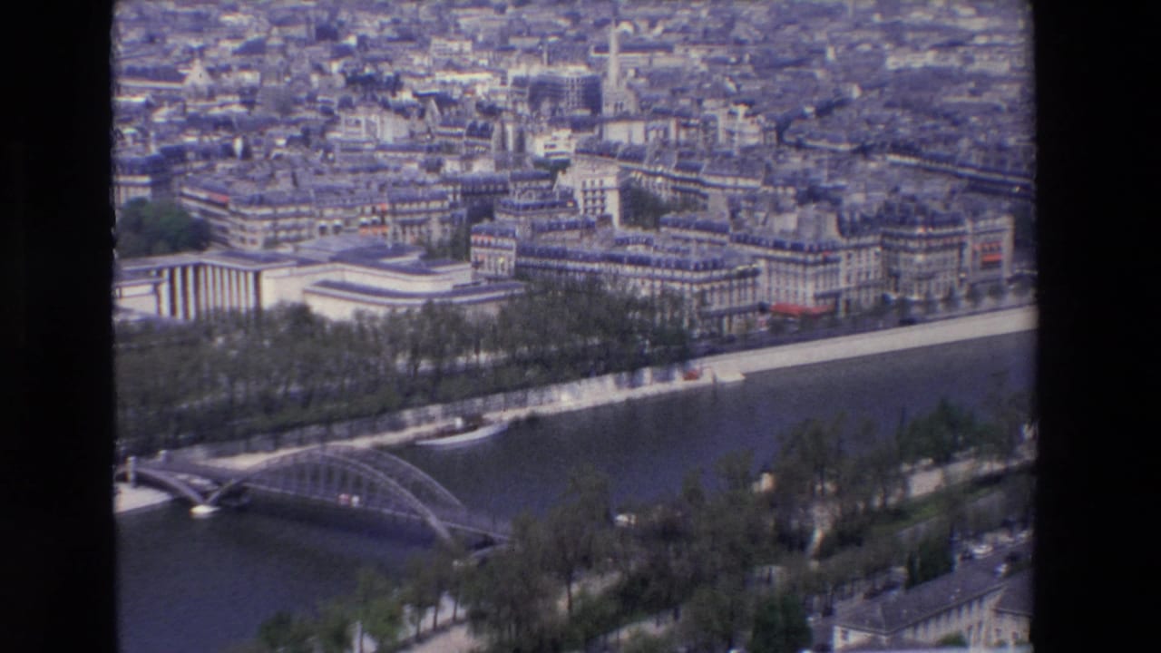 1969 aerial footage over Paris showing a metal bridge across the Seine, tree-lined quays, pale buildings, and dense urban blocks beyond the river.