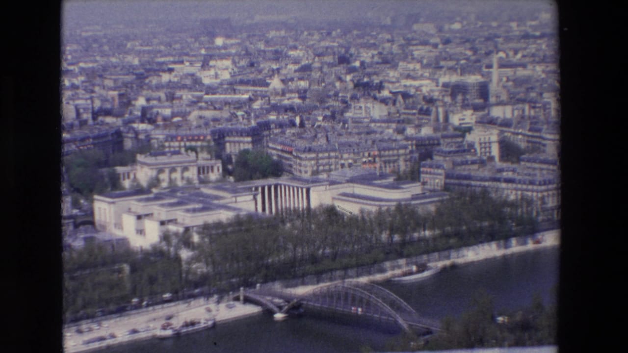 1969 8mm aerial view of Paris, showing pale buildings, the Seine embankment, a bridge, boats, and a hazy city background.