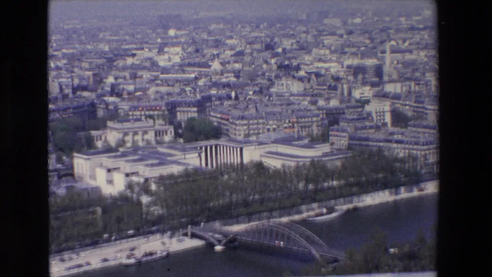 1969 8mm aerial view of Paris, showing pale buildings, the Seine embankment, a bridge, boats, and a hazy city background.