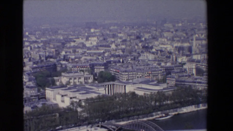 Aerial 1969 Paris frame with the Seine at the lower edge, a bridge, tree-lined embankment, pale civic buildings, and dense urban blocks beyond.
