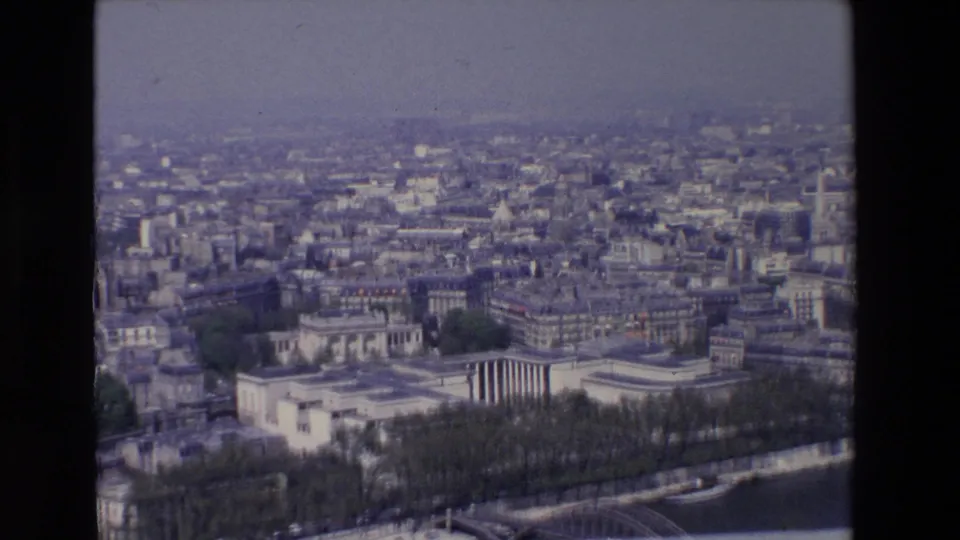 1969 8mm Paris aerial view with soft focus, showing rooftops, pale buildings, a riverside road, boats, trees, and bridge structure near the Seine.
