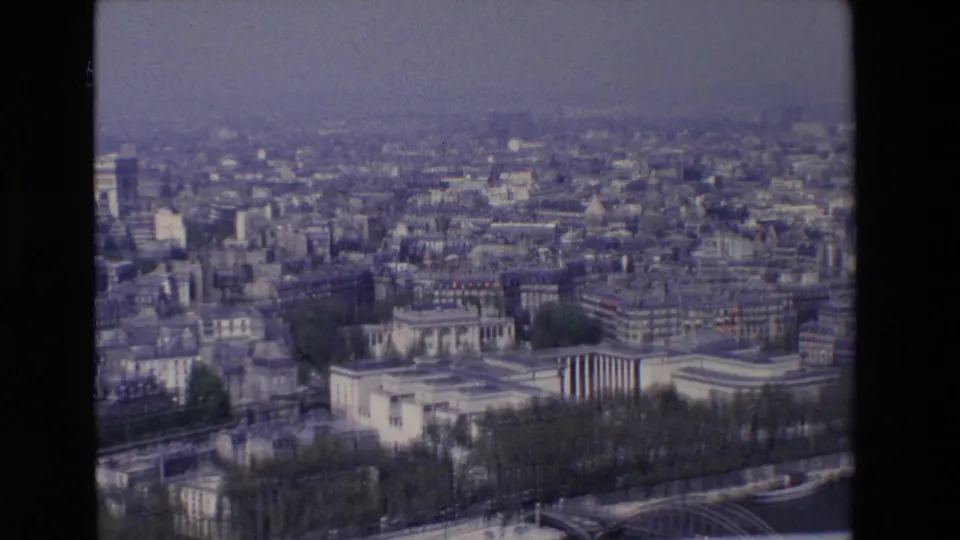 Late-1960s Paris aerial frame looking across the Seine riverfront toward pale buildings, dense city blocks, a bridge, trees, and a hazy skyline.