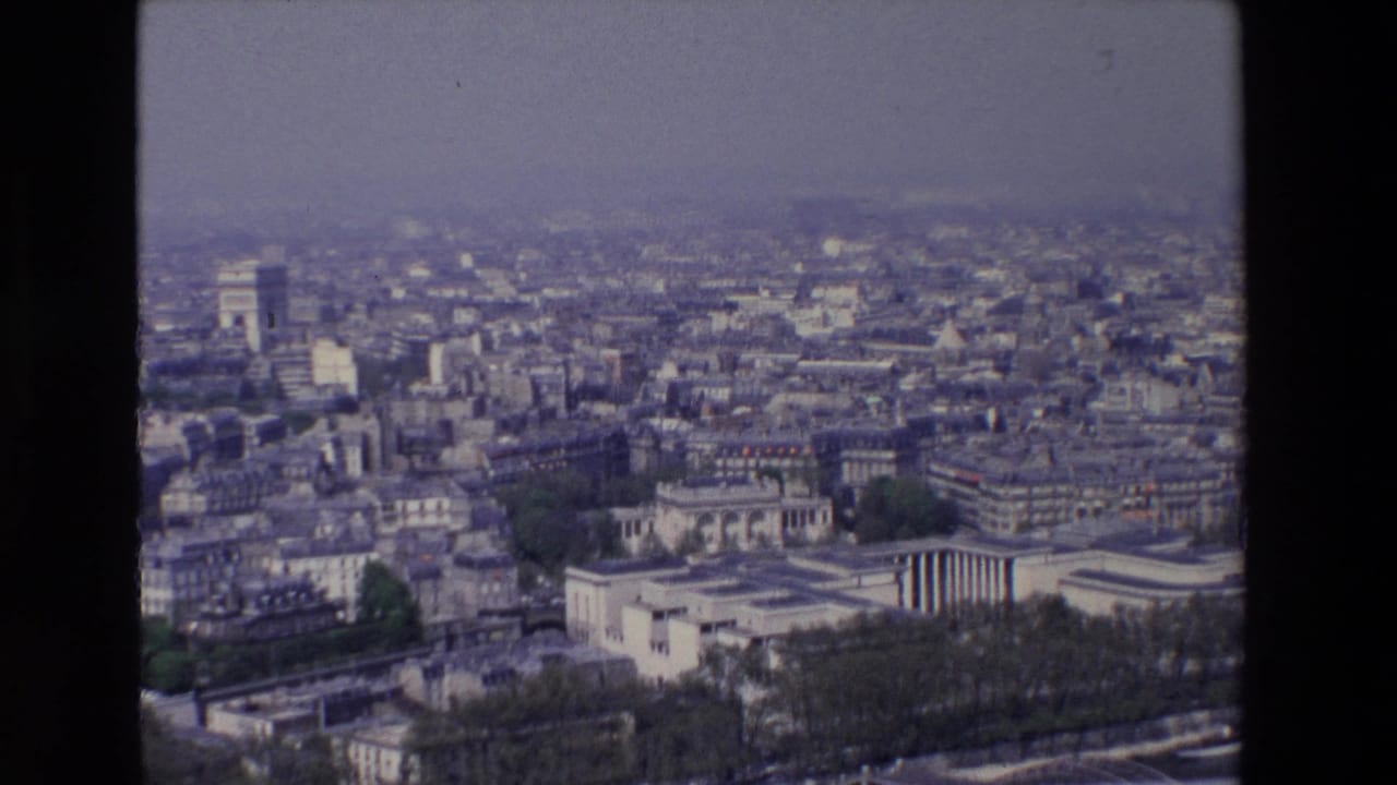 1969 8mm view over Paris rooftops and avenues, with the Arc de Triomphe visible in the distance beneath a hazy overcast sky.