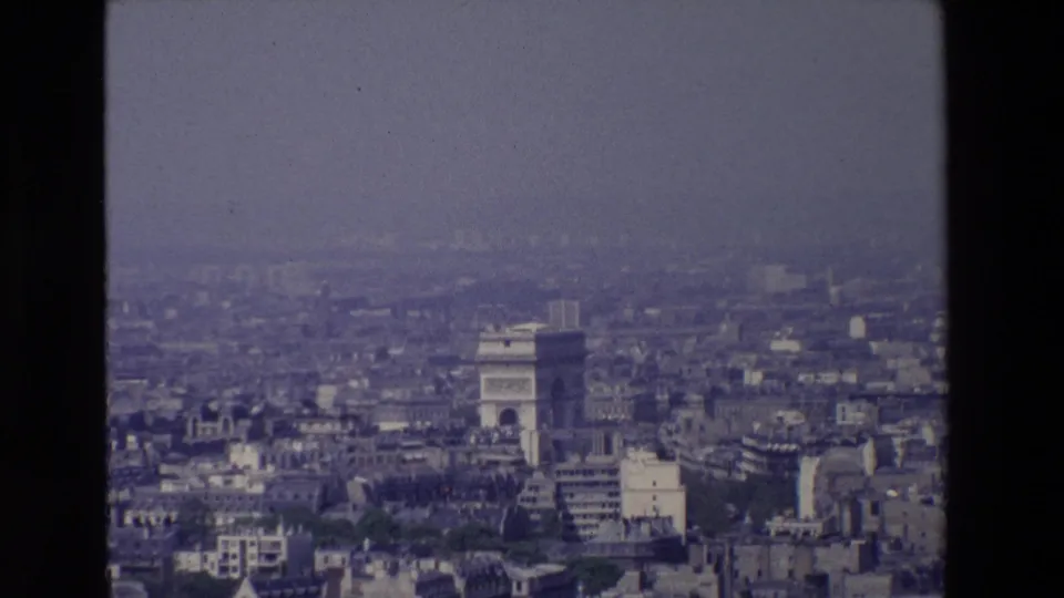 Late-1960s Paris 8mm frame with the Arc de Triomphe more prominent amid surrounding rooftops, modern blocks, and a hazy sky.