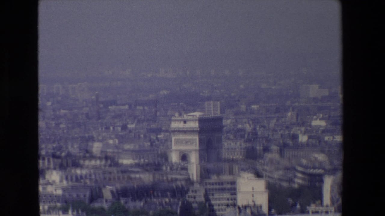 1969 Paris home-movie frame showing the Arc de Triomphe from a high aerial vantage point, surrounded by hazy rooftops and dark 8mm frame borders.