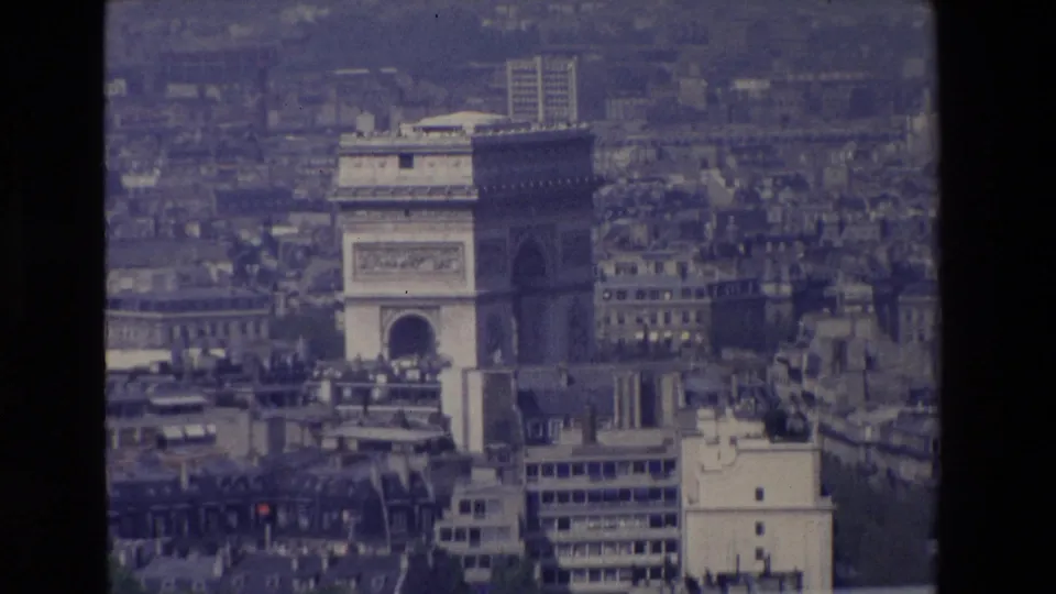 1969 Paris home-movie frame with a closer aerial view of the Arc de Triomphe, surrounding buildings, and visible grain in a hazy cityscape.