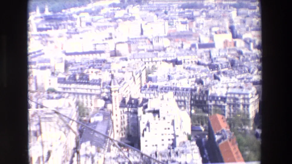 1969 aerial home-movie frame over Paris rooftops, with bright exposure washing parts of the cityscape and foreground structure visible at the lower edge.
