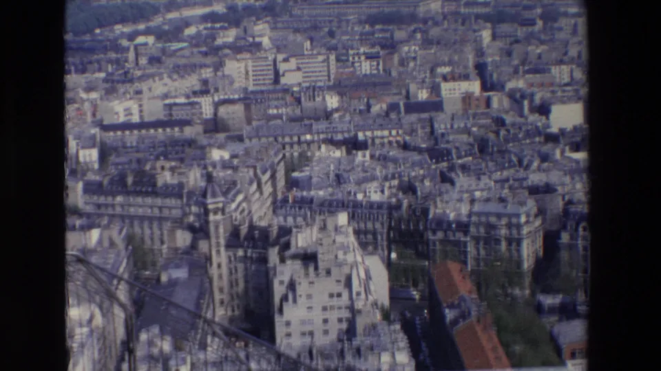 1969 Paris aerial frame looking across dense rooftops and avenues in softer blue light, with a foreground structure crossing the lower frame.