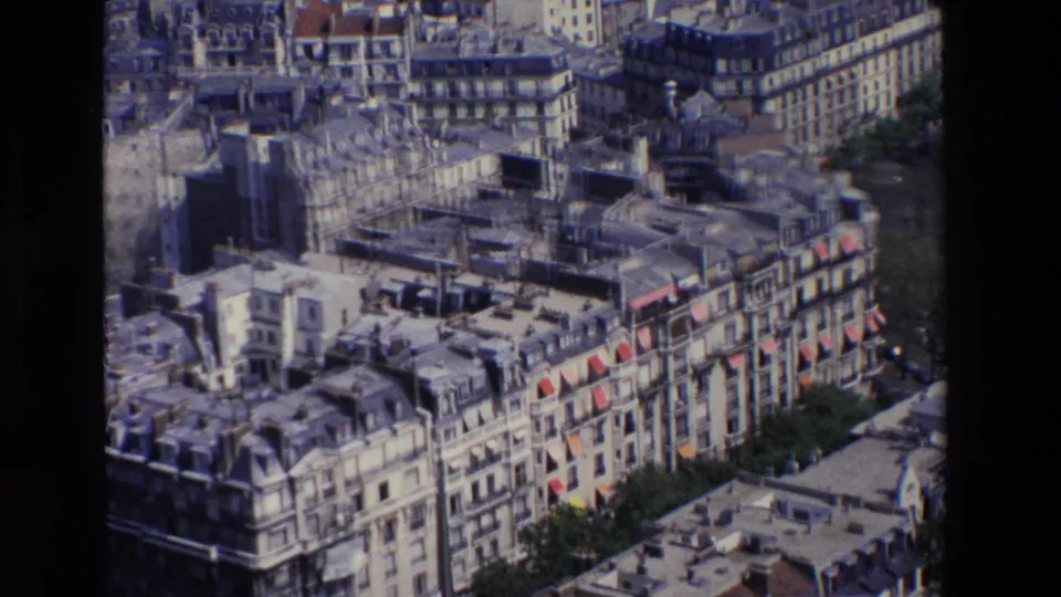 1969 Paris aerial frame centered on layered apartment roofs, pale stone facades, a taller dark building volume, street trees, and compact city blocks.