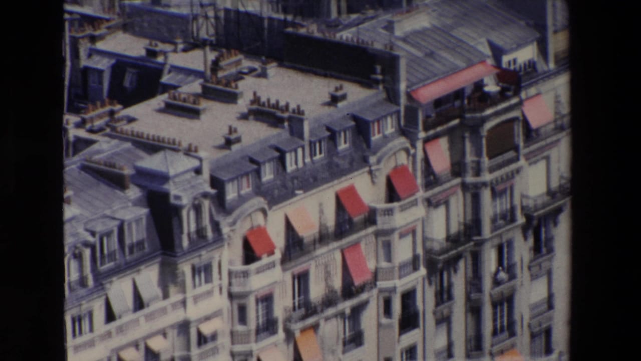 1969 Paris home-movie frame showing Haussmann-style apartment facades, gray mansard roofs, dormer windows, balconies, and red awnings from a high vantage point.