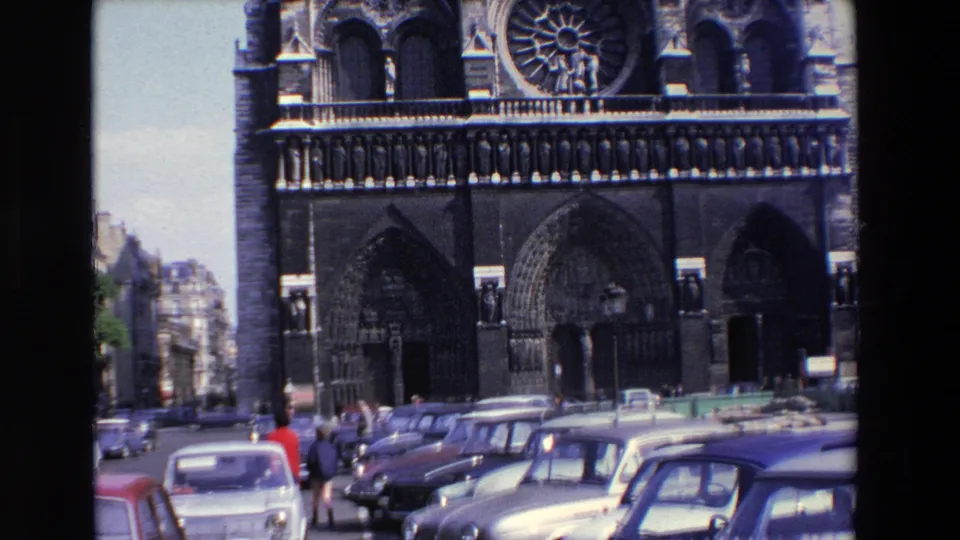 1969 Paris home-movie frame of Notre-Dame Cathedral with parked cars filling the lower foreground and small pedestrian figures near the entrances.