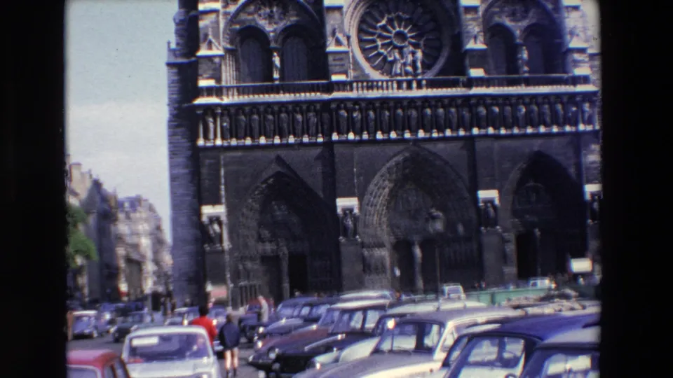 1969 Paris 8mm frame showing the cathedral facade, foreground automobiles, and pedestrians including a red-clad figure near the lower center-left.