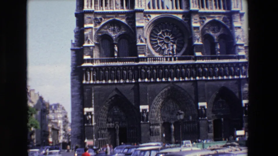 1969 Paris frame with Notre-Dame Cathedral centered behind street traffic, parked automobiles, and several pedestrians walking near the church frontage.
