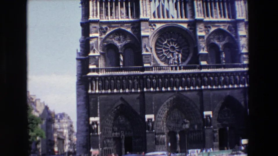 1969 8mm frame of Notre Dame Cathedral in Paris, showing the western facade, rose window, arched portals, sculptural stonework, and a narrow view of the street below.
