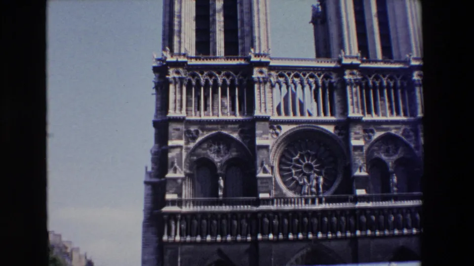 1969 8mm frame of Notre Dame Cathedral in Paris, angled upward toward the upper facade, rose window, tower columns, decorative arcades, and blue sky.