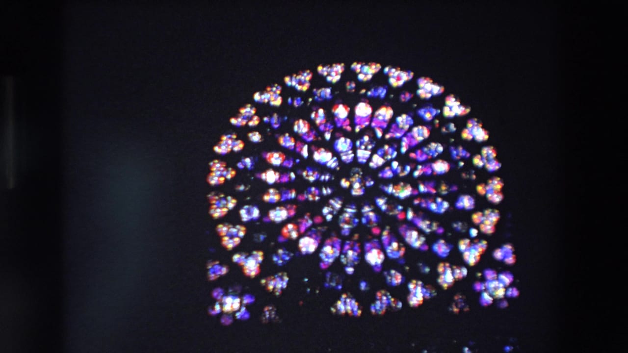 The camera holds on the luminous circular stained-glass rose window inside a dark Paris church, with saturated color and slight 8mm softness.