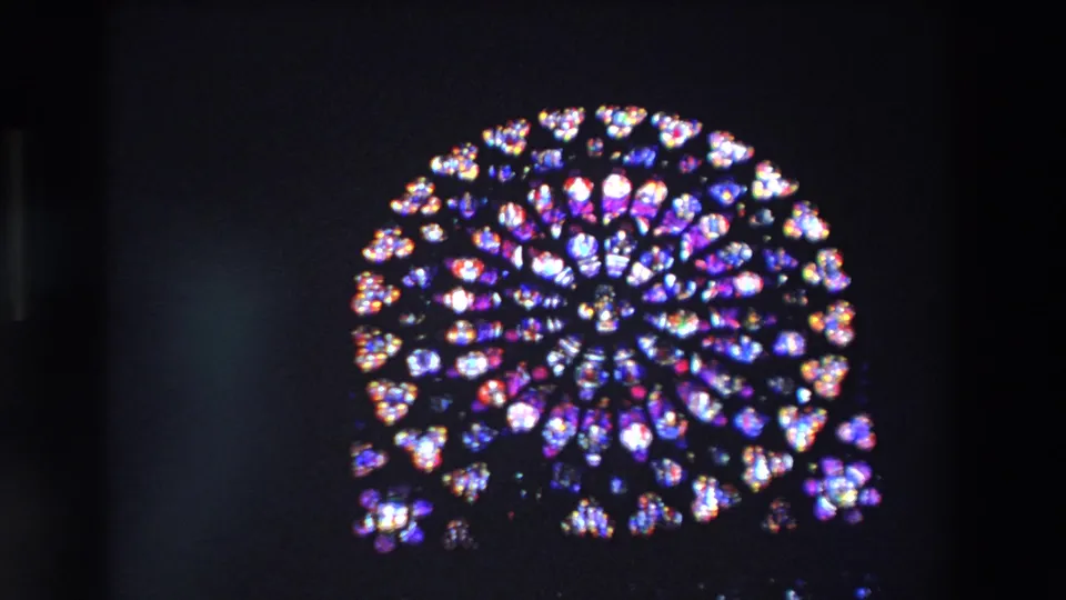 The camera holds on the luminous circular stained-glass rose window inside a dark Paris church, with saturated color and slight 8mm softness.