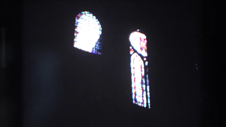Two arched stained-glass windows appear in the dark Paris church interior, one bright and distant and one taller panel glowing near the center.