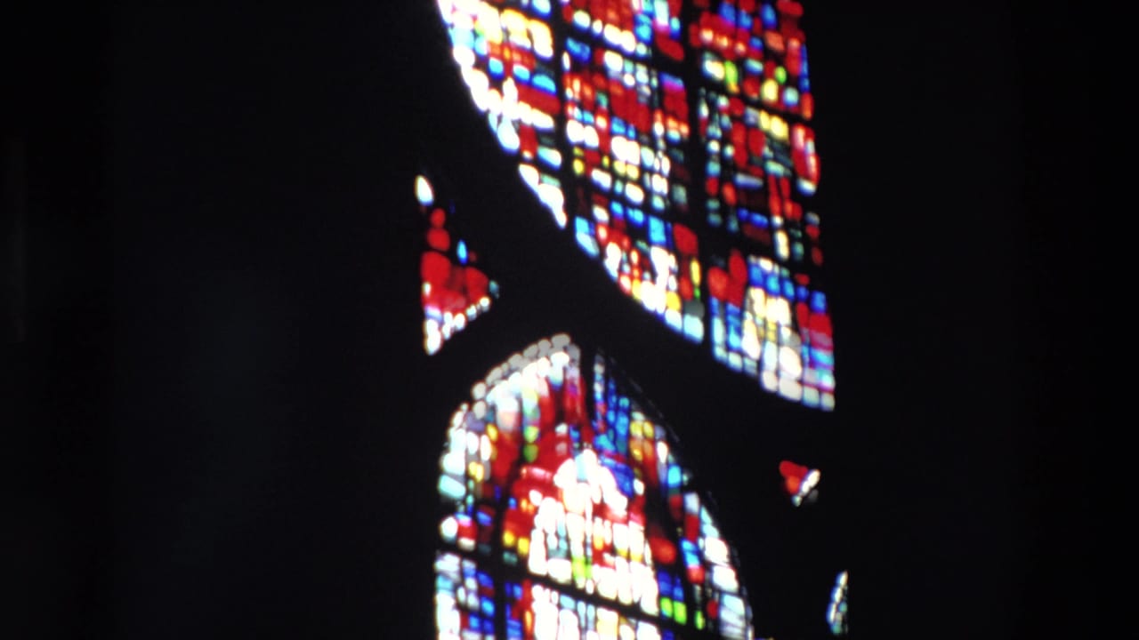 Paris church interior from 1969 home-movie footage showing tall stained-glass windows glowing red, blue, white, and yellow against deep shadow.