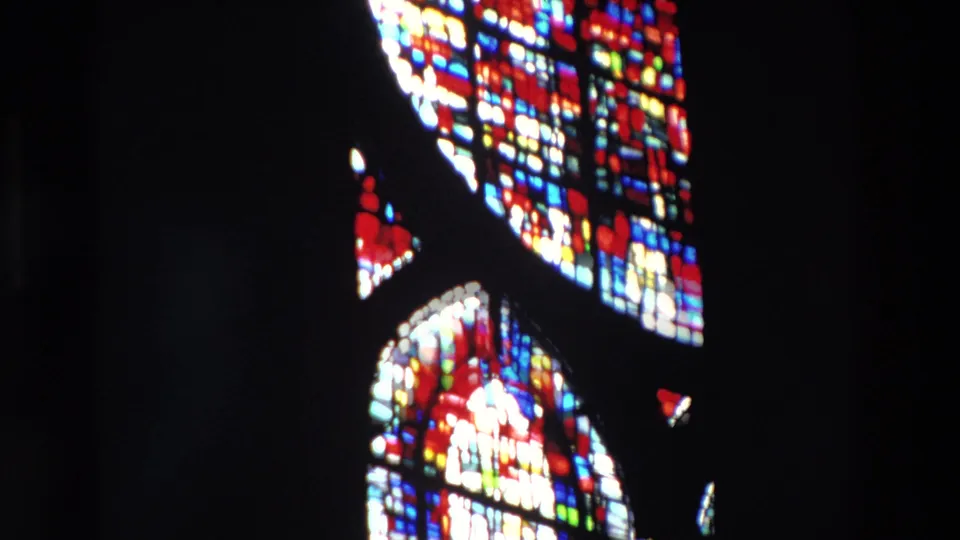 Paris church interior from 1969 home-movie footage showing tall stained-glass windows glowing red, blue, white, and yellow against deep shadow.