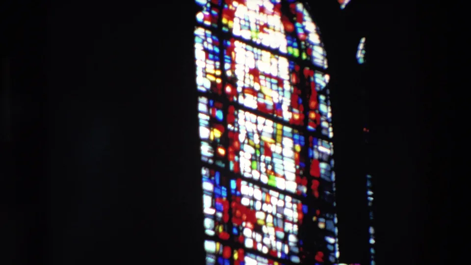 Close view of a stained-glass church window from the 1969 Paris footage, with colorful geometric leadwork and religious-looking forms softened by 8mm blur.