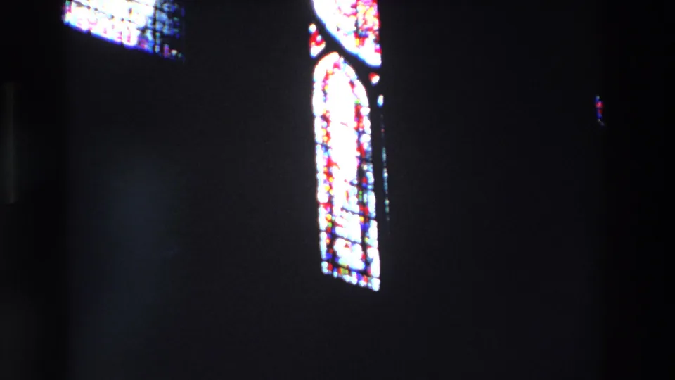 1969 Paris 8mm frame showing angled stained glass panes near the upper wall of a dark church interior, with bright colored sections and soft film grain.