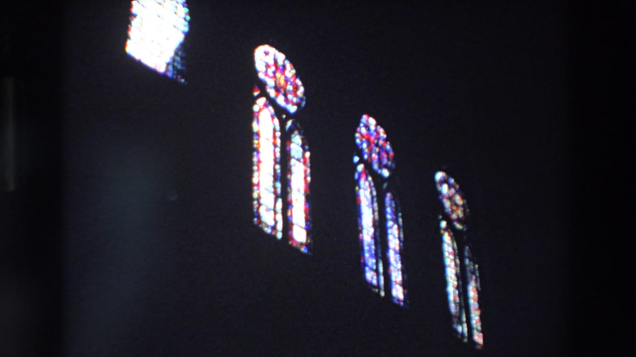 In 1969 Paris home-movie footage, several tall stained-glass windows glow red, blue, and white against a shadowed church interior.