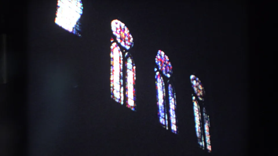 In 1969 Paris home-movie footage, several tall stained-glass windows glow red, blue, and white against a shadowed church interior.