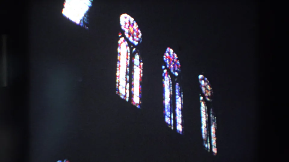 A handheld 8mm frame shows three narrow stained-glass window groups glowing inside a dark Paris church interior, with soft focus and visible film grain.