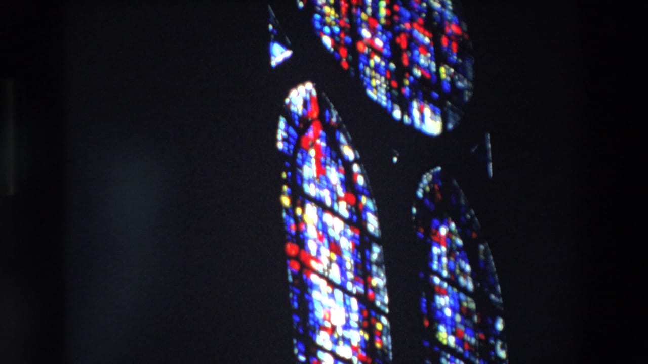 1969 Paris home-movie frame showing tall arched stained-glass church windows glowing blue, red, and yellow against a dark interior.