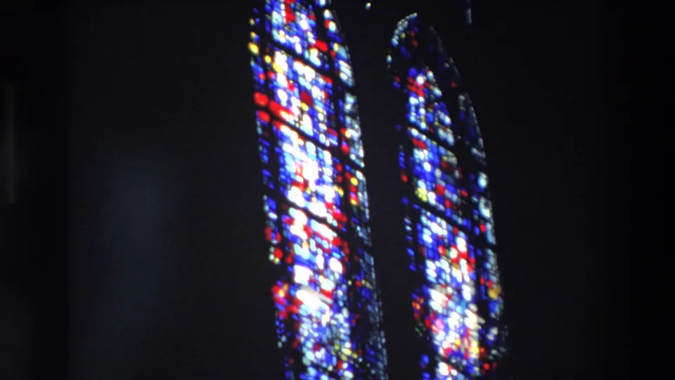 1969 Paris frame with bright vertical stained-glass panels, dominated by blue and white light with red details, inside a dark church space.