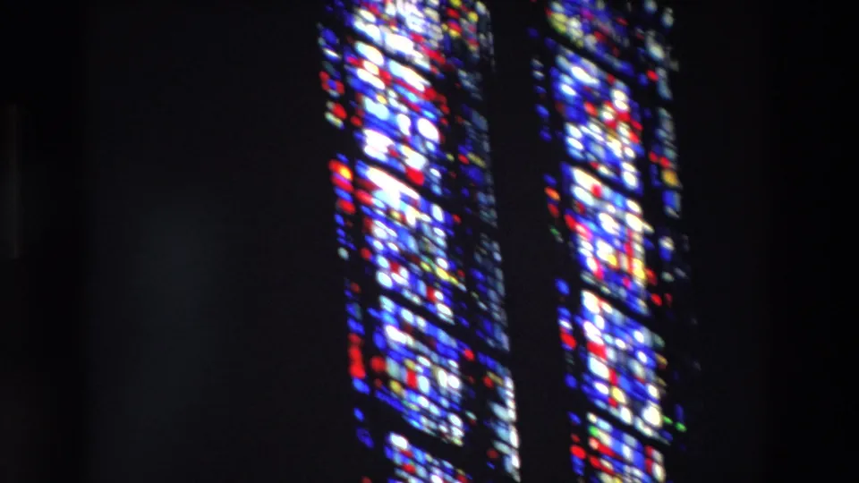 1969 Paris home-movie still of two narrow arched stained-glass windows glowing with blue, red, white, and yellow light in a dark church interior.