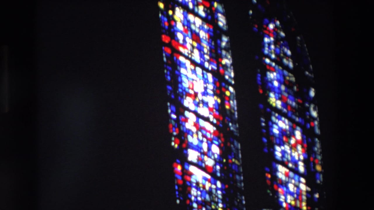 1969 Paris 8mm frame of tall stained glass panels glowing red, blue, white, and yellow against a dark church-like interior.