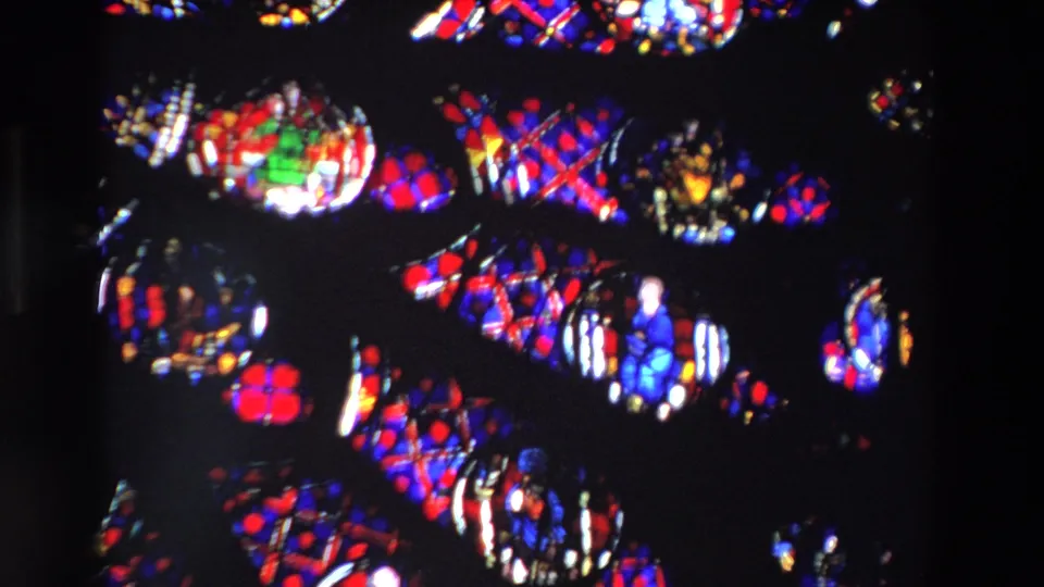 1969 Paris frame of stained glass with a blue-robed religious figure, circular medallions, and red geometric panels filmed from a handheld visitor viewpoint.