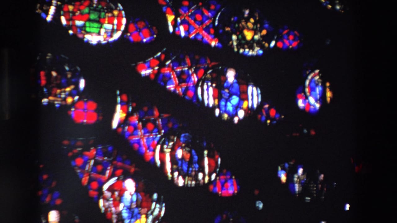 Close-up 1969 Paris home-movie frame of saturated red, blue, green, and yellow stained glass sections with religious figures and geometric leadwork in a dark church setting.