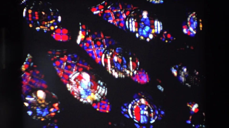 1969 Paris 8mm close-up of illuminated stained glass figures and patterned red and blue sections, framed tightly with surrounding darkness.