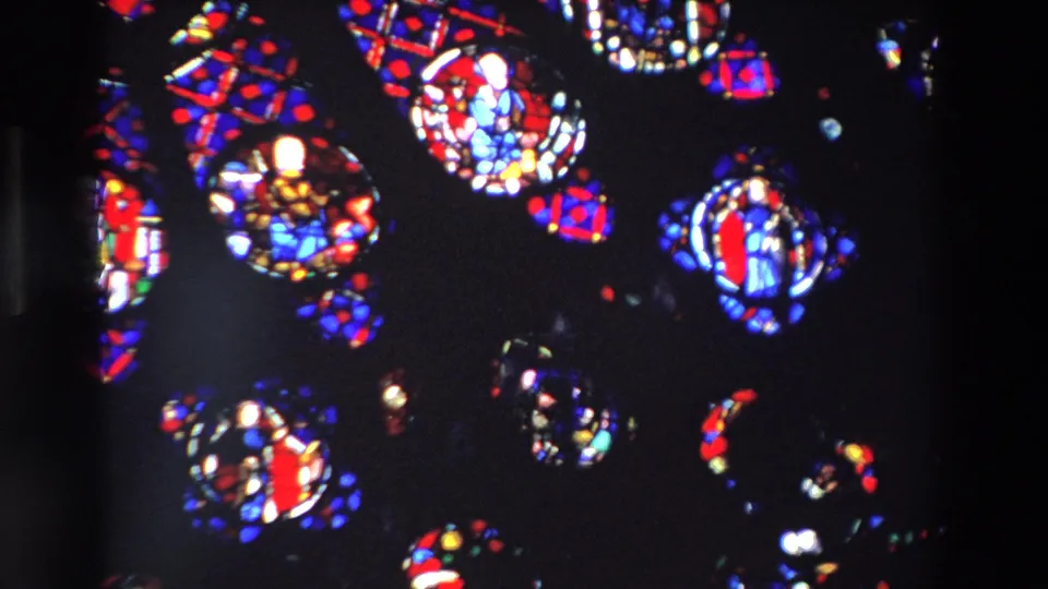 1969 Paris home-movie still of red, blue, and white stained glass details with religious figure motifs, softened by grain and low interior light.