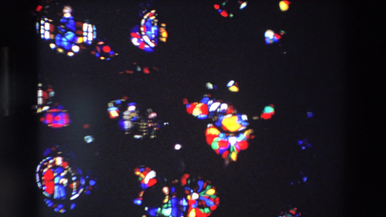 1969 Paris home-movie frame showing bright red, blue, green, and yellow stained glass fragments against a dark cathedral interior.