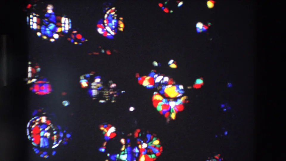 1969 Paris home-movie frame showing bright red, blue, green, and yellow stained glass fragments against a dark cathedral interior.