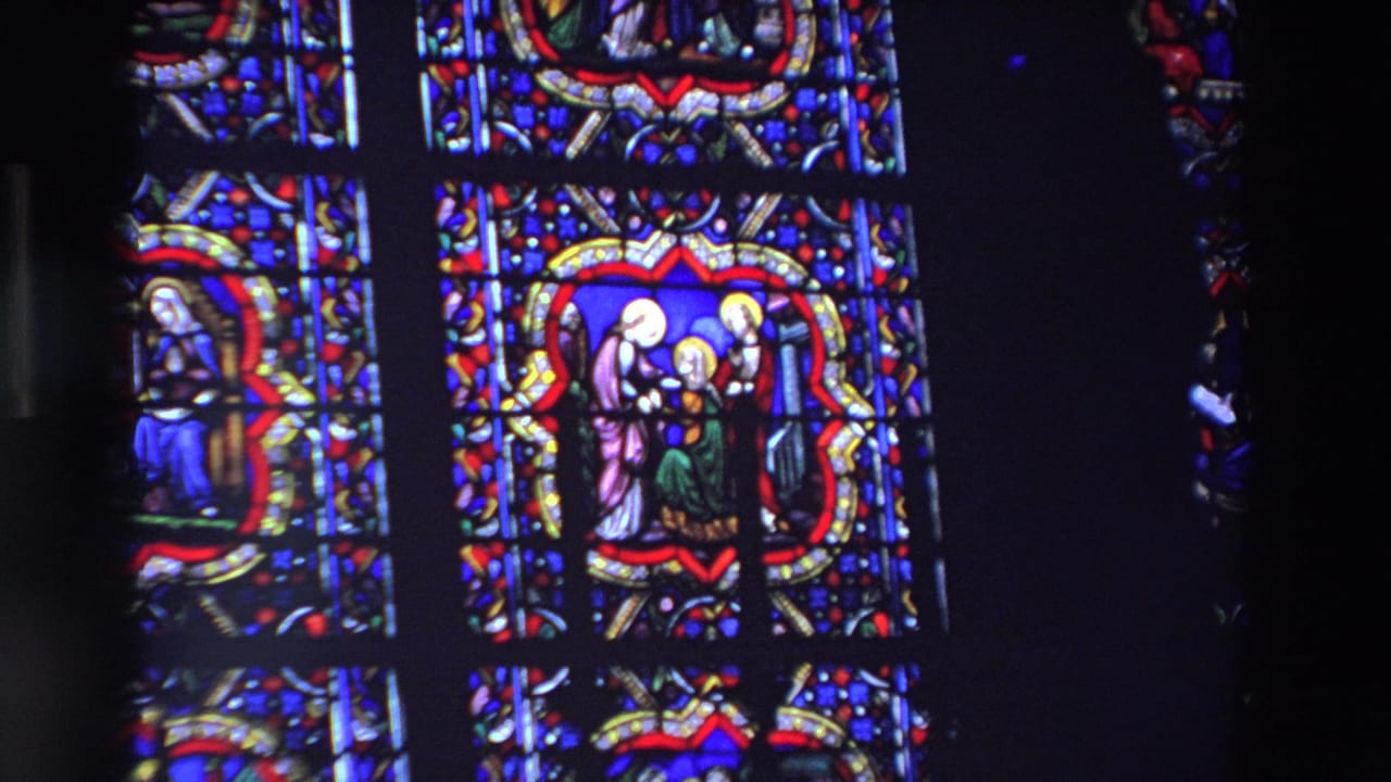 Wide 1969 Paris 8mm view of tall stained glass panels with blue, red, and gold religious scenes inside a dark church interior.