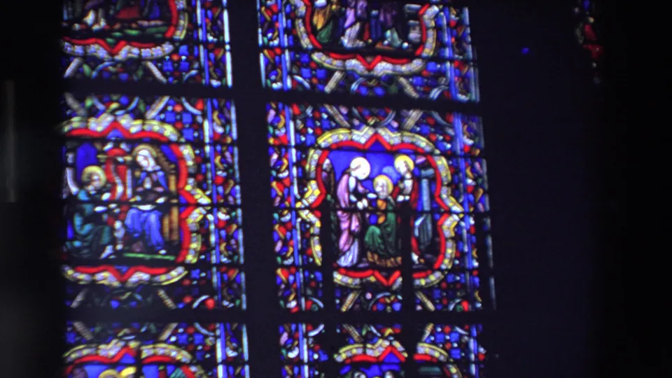 Two sections of stained glass in a Paris church show haloed religious figures, patterned borders, and deep blue glass filmed from a visitor perspective.
