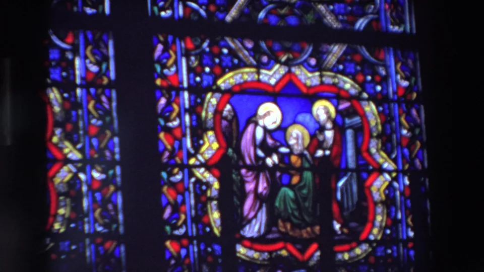 Closer 8mm view of a stained glass panel in a dark Paris church, showing haloed religious figures within a red and gold border.